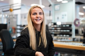 Gry Amalie Rosberg at her work station at Hatteland Technology's production facility in Aksdal, Norway, where she assembles maritime monitors and maritime displays. She is smiling, facing the camera, wearing a black sweater. Behind her is various technical equipment. 