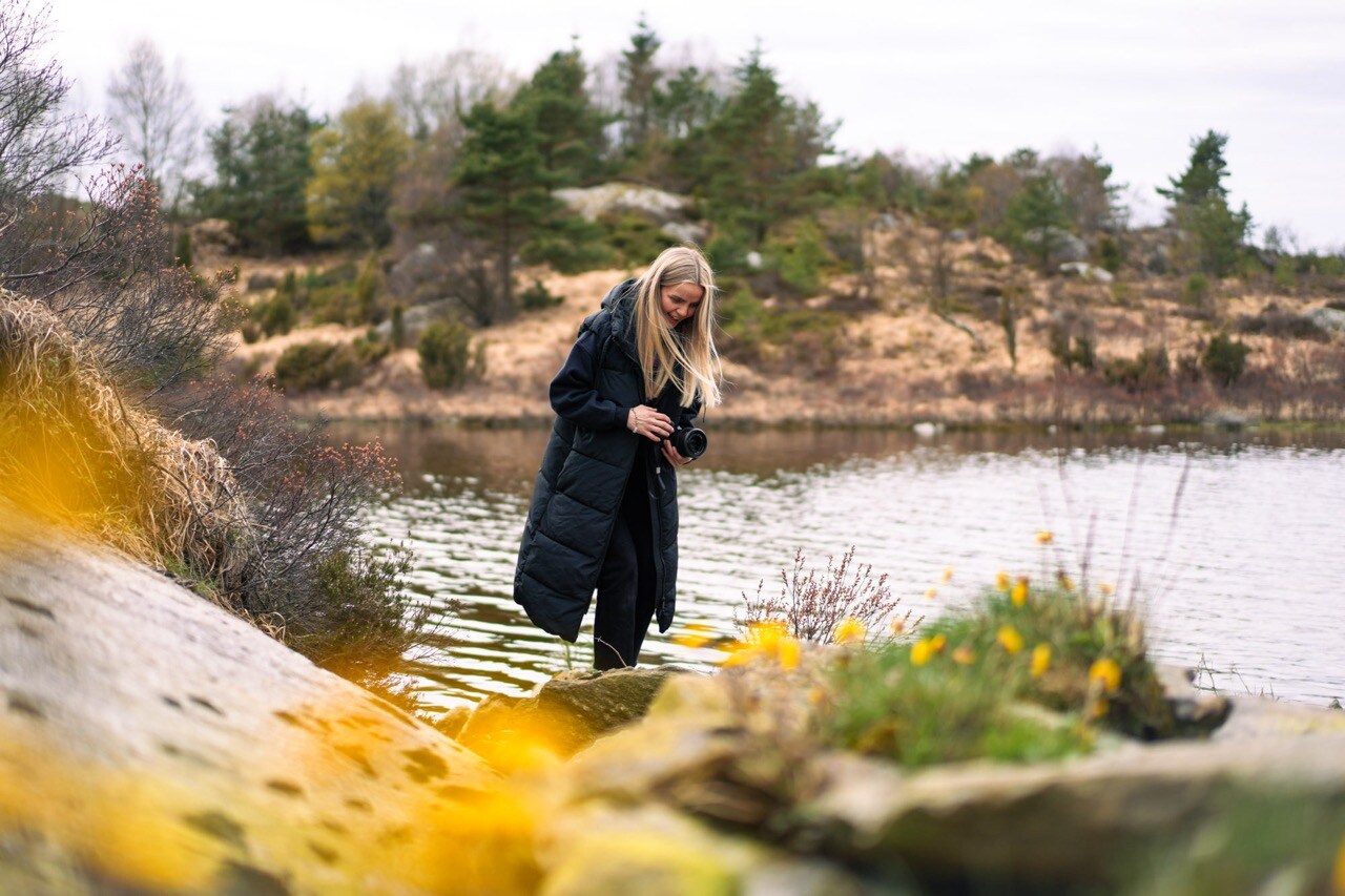 Gry Amalie Rosberg is in the center of the photo, holding a camera. In the foreground, yellow flowers, in the background, Aksdalsvatnet. She is wearing a dark winter coat.