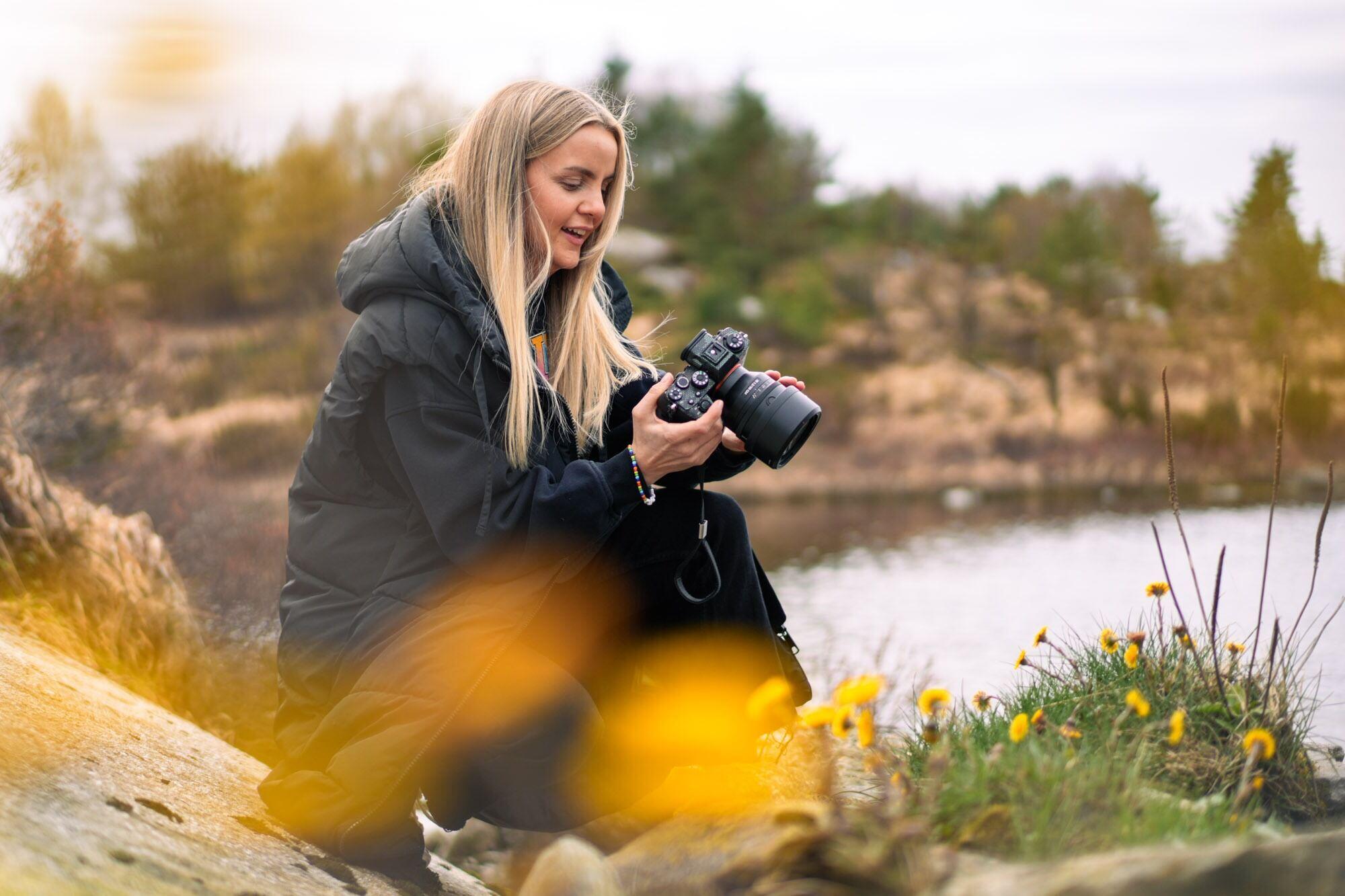 Gry Amalie Rosberg is in the center of the photo, kneeling, looking at the LCD screen of her camera. She is photographing wildflowers on the shore of Aksdalsvatnet. In the foreground, yellow flowers, in the background, a lake and surrounding landscape. Gry Amalie is wearing a dark winter coat.