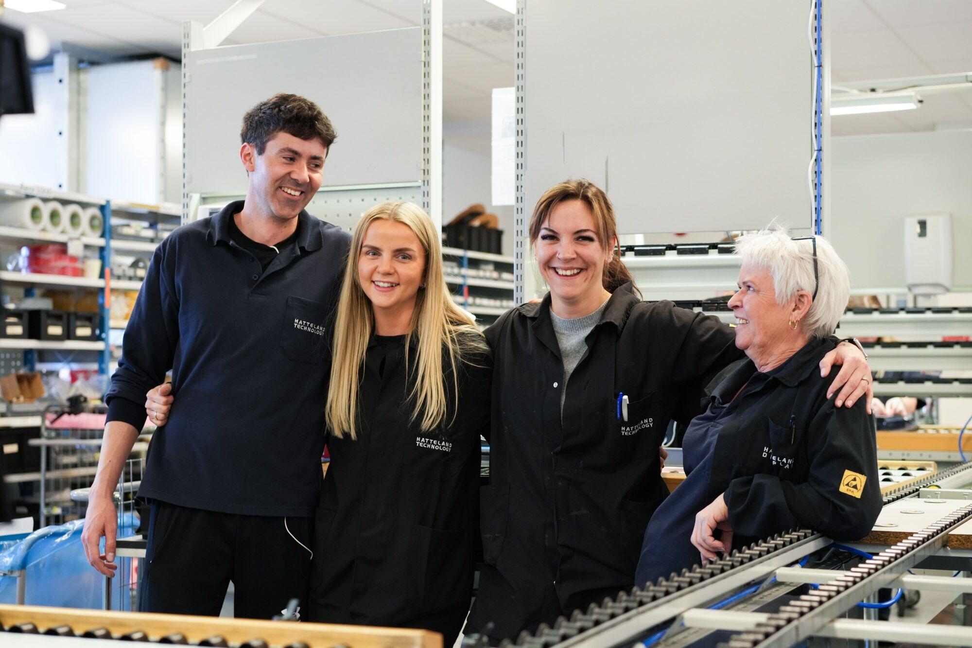 This is a picture of Gry Amalie and some of her colleagues at Hatteland Technology's production facility in Aksdal, Norway, where the manufacture maritime displays, monitors, and industrial IT hardware. They are huddled together, laughing. They are wearing dark Hatteland Technology workwear.
