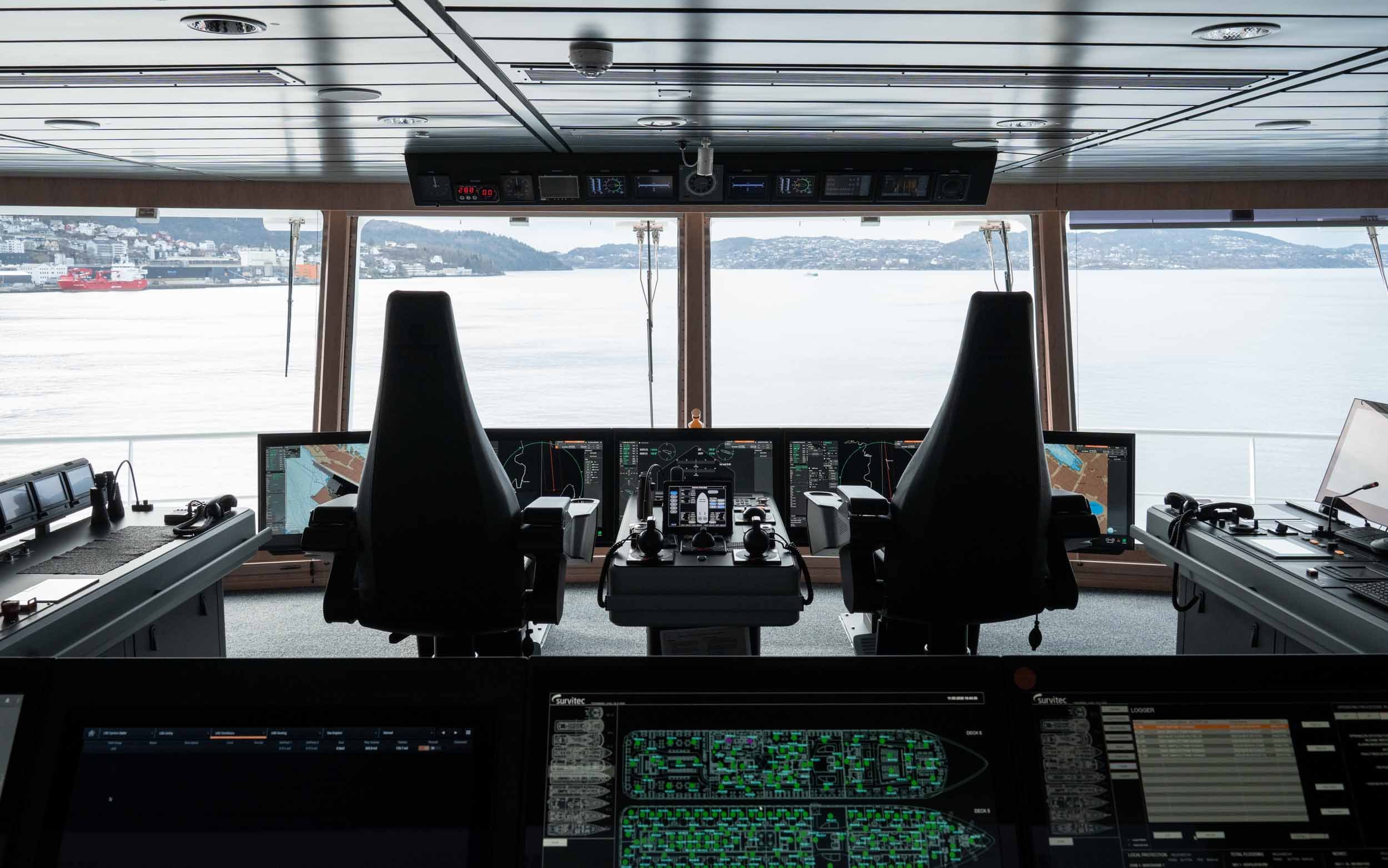 The forward view from the bridge on board Havila Polaris. In the lower half of the picture, an array of Hatteland Technology monitors can be see. Outside the window is the fjord. It is a cloudy, but calm day. 