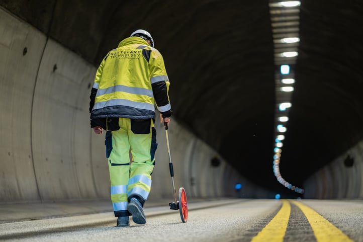 Morten Smestad from Hatteland Technology is surveying a road tunnel for a surveillance solution. His in the left part of the photo, wearing a yellow, reflective suit, and a white helmet. The tunnel is dark, but lit by lamps. 