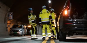 Three engineers discuss a surveillance solution in a road tunnel in Norway. They are wearing yellow, reflective work attire, helmets. The tunnel is dark. Two yellow centre lines run through the middle of the picture.