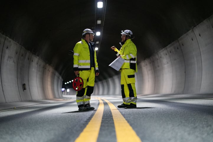 Morten Smestad and Jan Olav Larssen from Hatteland Technology, surveying a road tunnel for a surveillance solution. They're in the center of the photo wearing yellow, reflective suits, and white helmets. The tunnel is dark, but lit by lamps. 