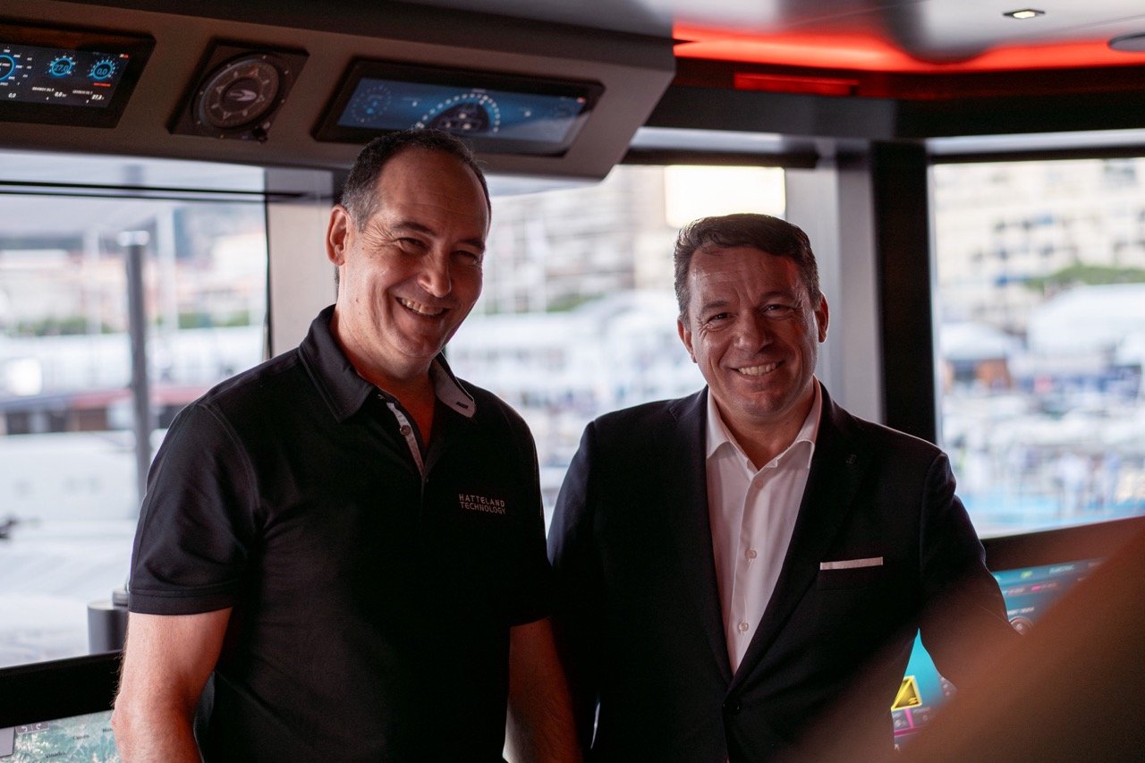 The picture shows Mehdi Bounoua, sales director EMEA at Hatteland Technology, and Massimo Bugli, general manager of Furuno Italia, together on the bridge of superyacht Baglietto T52 at the Monaco Yacht Show 2023. The gentlemen are both smiling, facing the camera. In the background the bridge system of the yacht is partly visible. In the distance you can see a blurred-out bay of Monaco. 