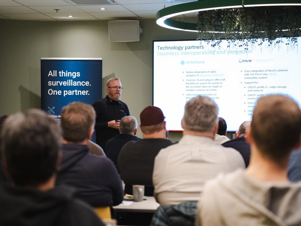 Bildet viser John Ørnebjerg fra Bosch, som presenterer under Bosch/Milestone-samling i Oslo. I forgrunnen sitter det en interessert forsamling. Bak John er et Hatteland Technology-banner. 