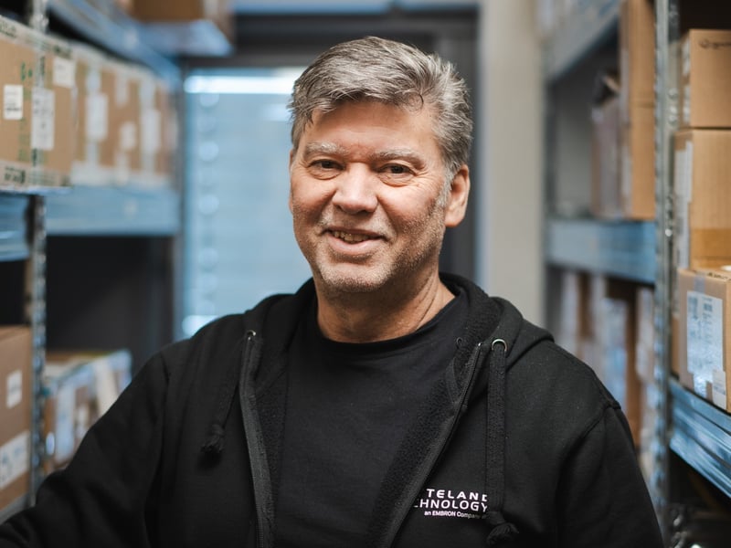 A close-up of team leader Fernando Oliveira at Hatteland Technology's warehouse. He is wearing a black hoodie and is smiling. In the background there are cardboard boxes on shelves.