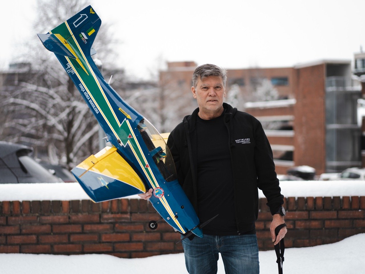 Fernando Oliveira is holding an RC airplane similar. They are surrounded by office buildings covered in snow, as it's a winter's day. Fernando is wearing jeans and a black hoodie, facing the camera. He is holding a remote controller. 