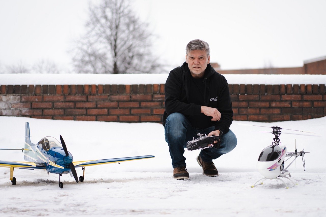 Fernando Oliveira is squatting next to two of his RC aircraft, an airplane in the left of the picture and a helicopter on the right side. They are standing on the snow, as it's a winter's day. Fernando is wearing jeans and a black hoodie, facing the camera. He is holding a remote controller. 
