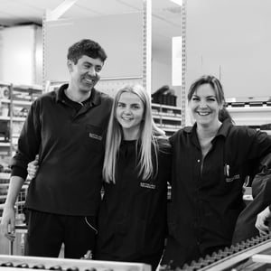 A group of Hatteland Technology workers at the production facility in Aksdal, Norway. From the left, a tall man with dark hair, then a blonde woman, and on the right a dark-haired woman. They are smiling.