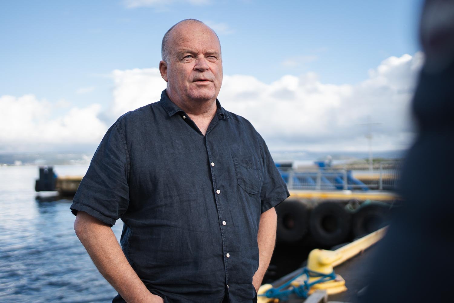 The picture shows Anders Bjerketveit, maritime CCTV expert and Senior Key Account Manager at Hatteland Technology. He is wearing a buttoned t-shirt. His hands are in his pockets. In the background you can see the sea and a ferry pier. The sky is blue.