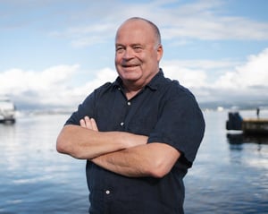 The image shows Anders Bjerketveit, Hatteland Technology's maritime CCTV expert. He stands on the dock, facing the camera, smiling. His arms are crossed. He is wearing a dark blue buttoned t-shirt. Behind him is the calm sea and blue skies.