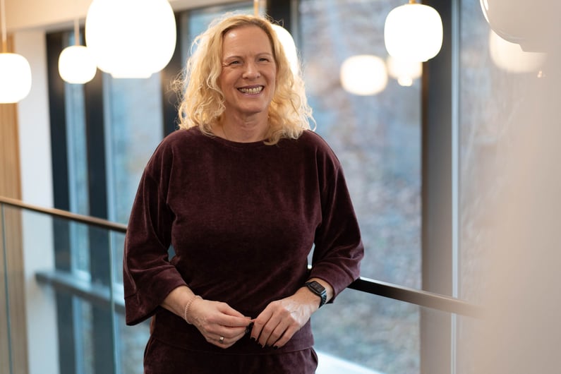 This is a photo of Elke Freisens, our office manager in Germany. She is standing in an office building, smiling. She is wearing a burgundy top. Behind her, there are spherical lamps and large windows.