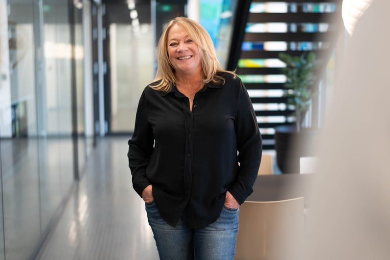 The picture shows Donna Pallonetti, office manager for our US branch, at Hatteland Technology's office in Lysaker, Norway. She is smiling. She is wearing a black shirt. Her hands are in her pockets. 