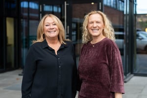 The picture shows Donna Pallonetti and Elke Freisens, office managers for Hatteland Technology in the US and in Germany. They are facing the camera, smiling. There is an office building behind them. 