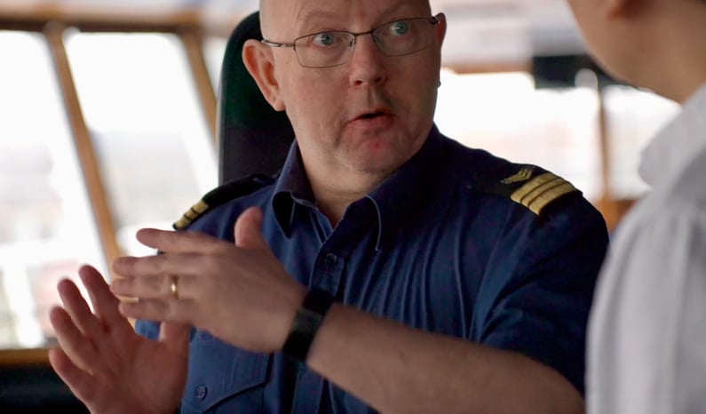 This is a picture of electronics engineer Knut-Olav Reistad on the bridge of MS Color Magic. He is wearing a blue mariner's shirt with shoulder/rank marks. He is wearing glasses. 