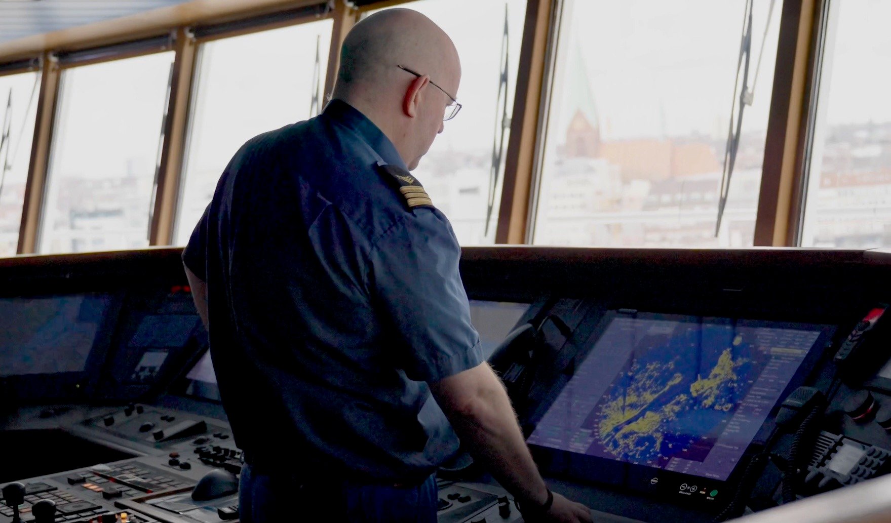 Here you can see electronics engineer Knut-Olav Reistad operate one of Hatteland Technology's marine monitors on board the MS Color Magic. There are several other Hatteland Technology monitors around him. He is wearing a blue mariner's t-shirt with shoulder marks. In the background, an array of large bridge windows. 