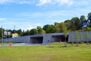 The picture depicts the opening and exit of Frodeåstunnelen, a road tunnel in Vestfold, Norway, for which Hatteland Technology supplied the AID solution based on the Sprinx(R) technology. The tunnel is surrounded by a green field and a green hillside. 