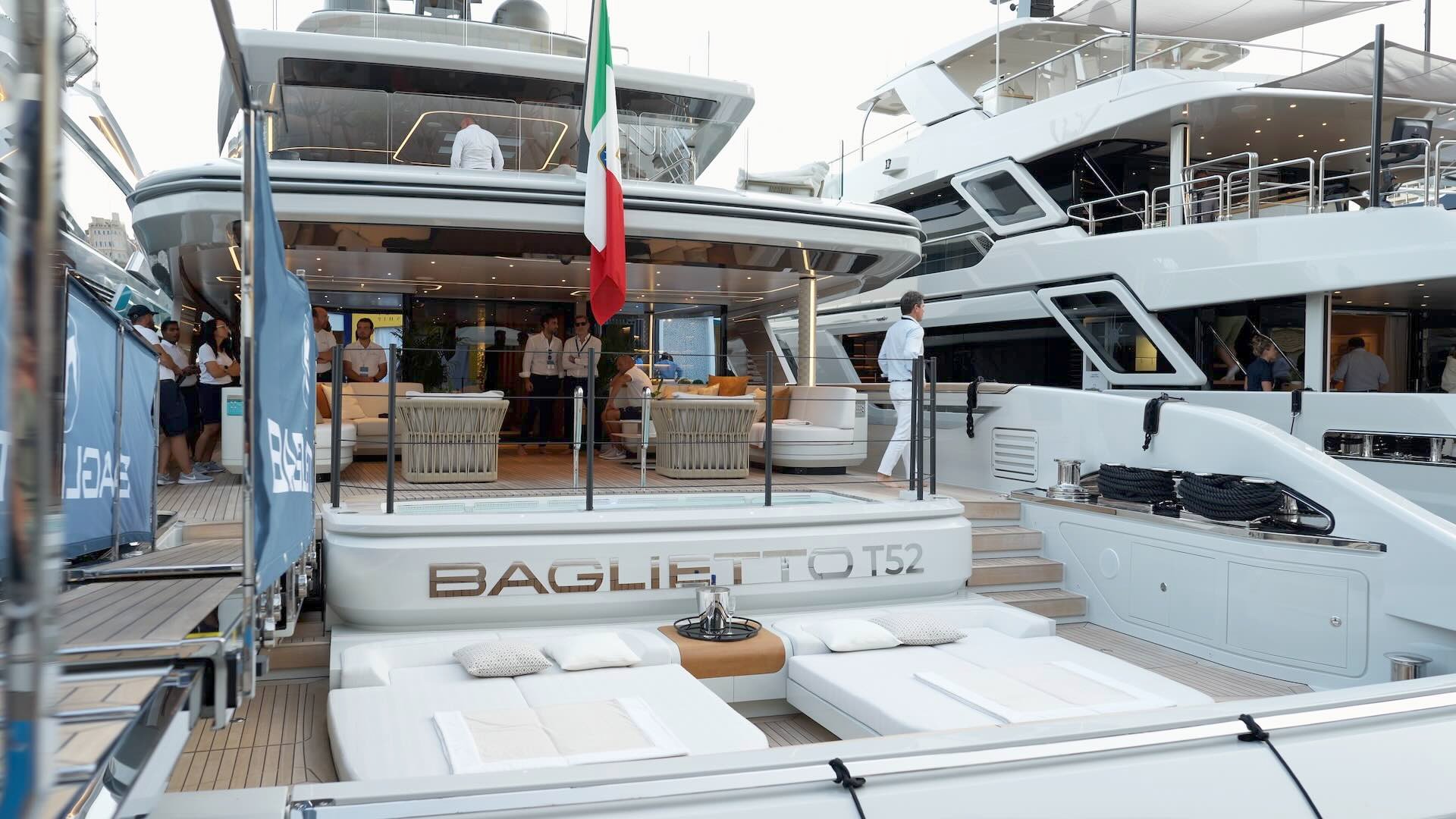 The image shows superyacht Baglietto T52 at the Monaco Yacht Show 2023. The yacht is seen from the rear, with an Italian flag visible. The crew is dressed in white shirts. 