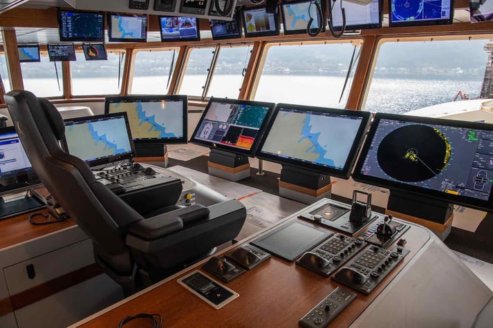 An internal image of a modern ship bridge with plenty of digital navigation equipment, such as monitors, thrusters and controllers. The captain's chair sits centrally in the image.