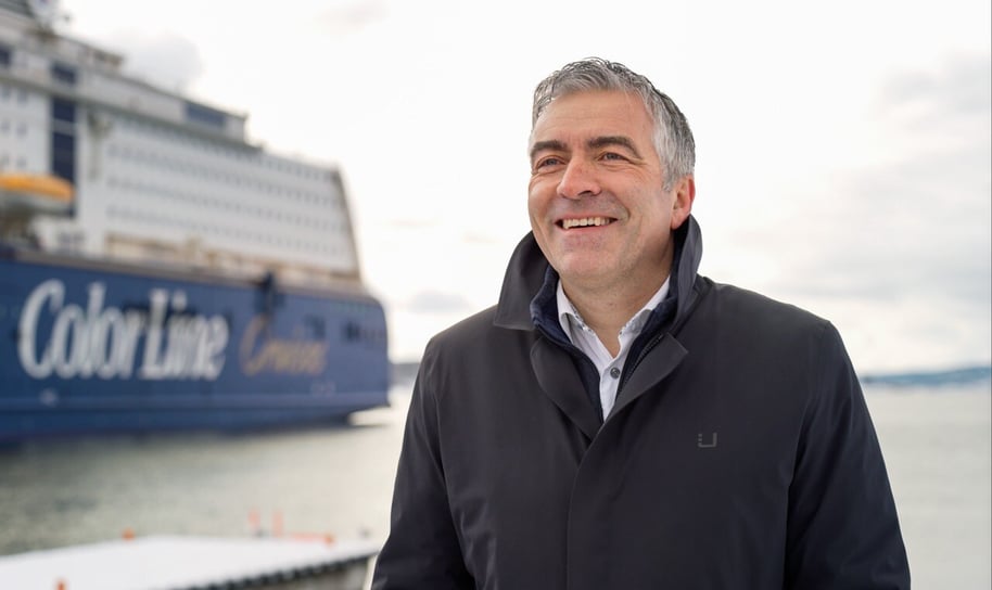 The CEO of Hatteland Technology, Lars-Skjelbred Eriksen, is standing in front of the MS Color Fantasy laying in port in Oslo harbor. It is a winter day, slightly overcast. He is wearing a black coat and a white shirt. He is smiling. The water is calm. The Color Line Cruises logo is visible on the ship's side.