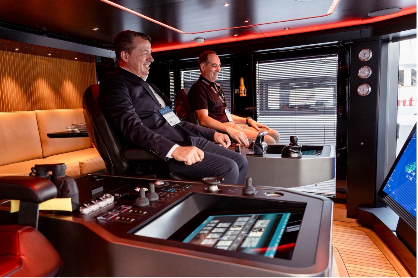 The image shows Massimo Bugli from FURUNO Italia, and Mehdi Bounoua from Hatteland Technology, in the wheelhouse of a yacht at the Monaco Yacht Show 2023. They are seated in the officers' chairs, smiling, exploring the bridge system.