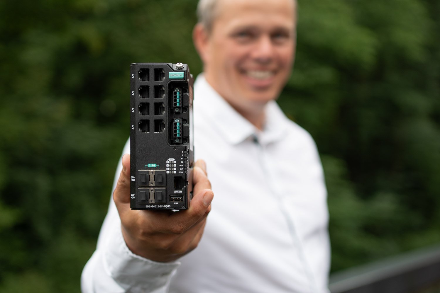 Mr. Gøran Labrå of Hatteland Technology holds up the Moxa EDS-G4012 to the camera. The unit is in focus, while Mr. Labrå is blurred out. He is smiling and wearing a white shirt. The background is made up of green trees.