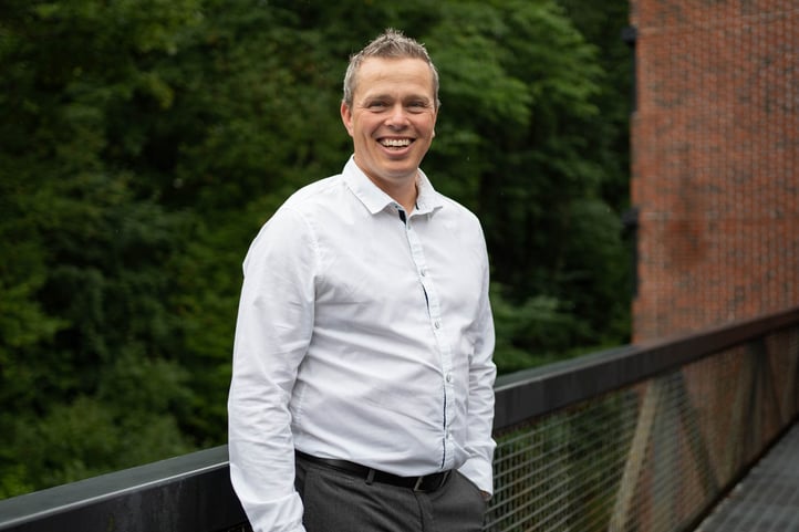The picture shows Mr. Gøran Labrå, Business development director at Hatteland Technology. He is wearing a white shirt, one hand in his pocket, with a big smile. There are green trees and an office building in the background.