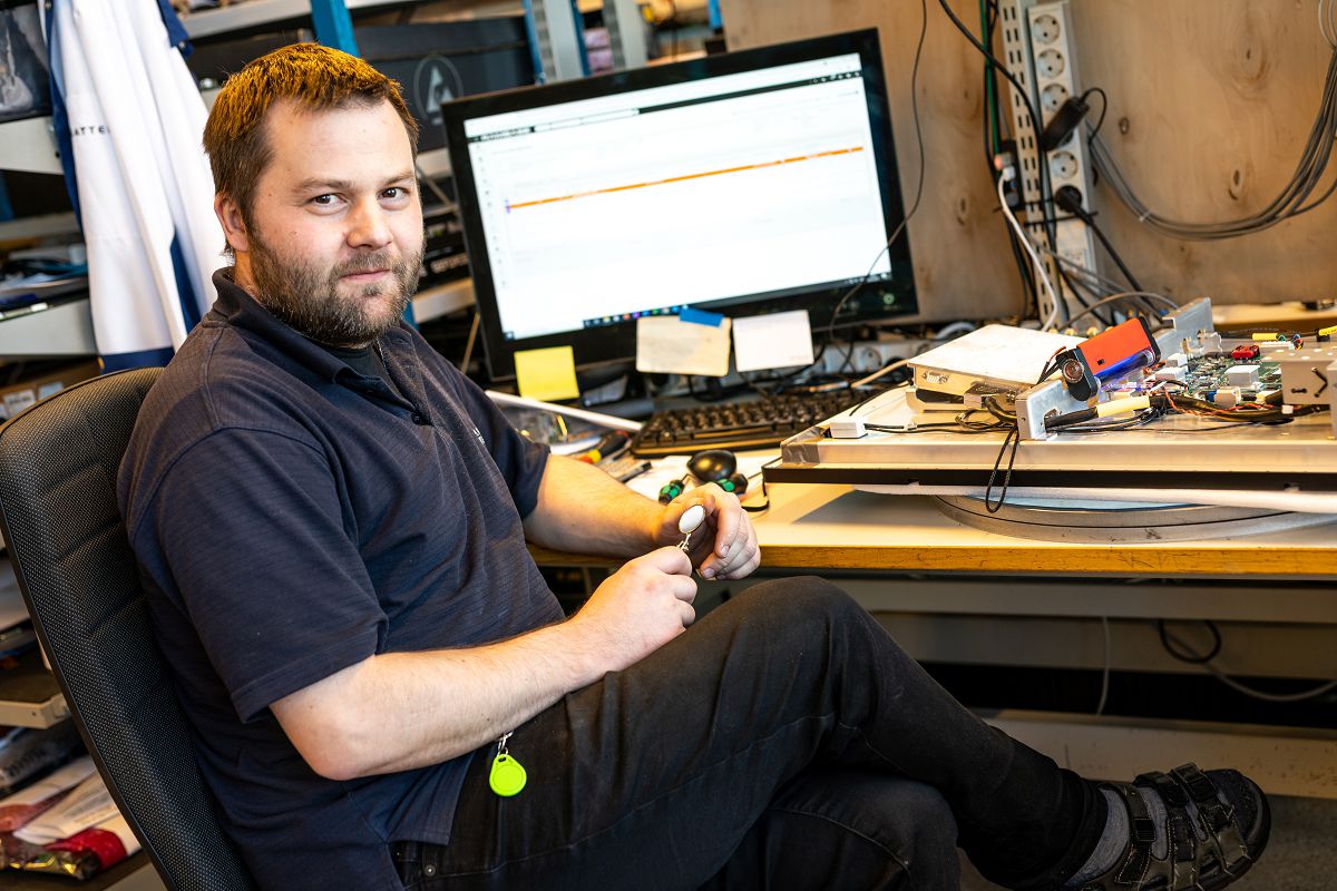 A worker at Hatteland Technology is sitting down in front of a computer. He is holding something in his hands, and is facing the camera. He has short hair and a beard.