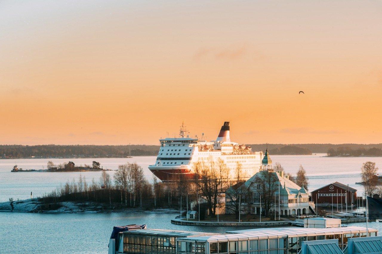 A modern passenger ferry is sailing on a calm winter's day in a Nordic fjord. The ferry is red and white. In the foreground, a few buildings and trees. It's a sunny day. 