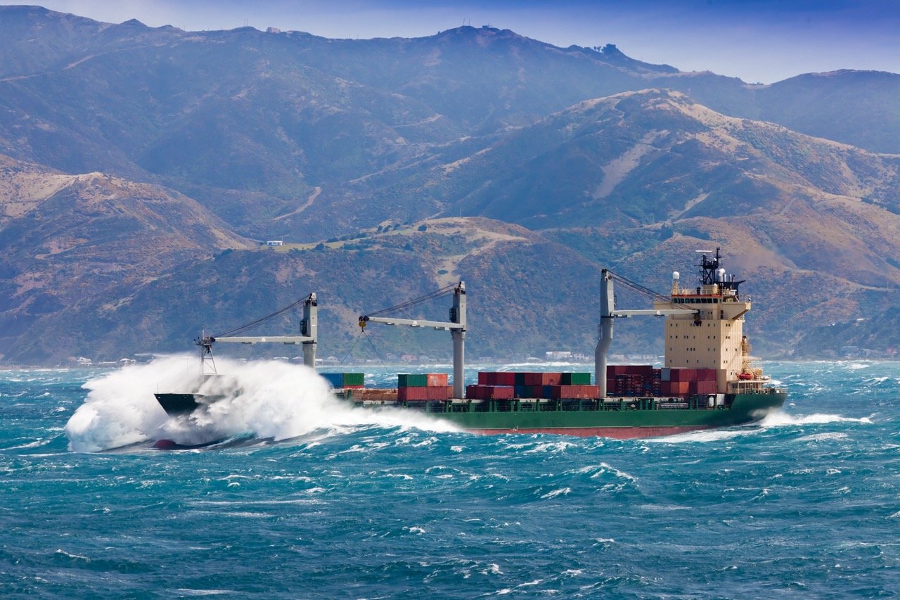 This is a picture of a green and red cargo ship sailing through rough seas. Waves are crashing over the ship's bow. In the background, a hilly landscape. It's a sunny, but windy, day. 