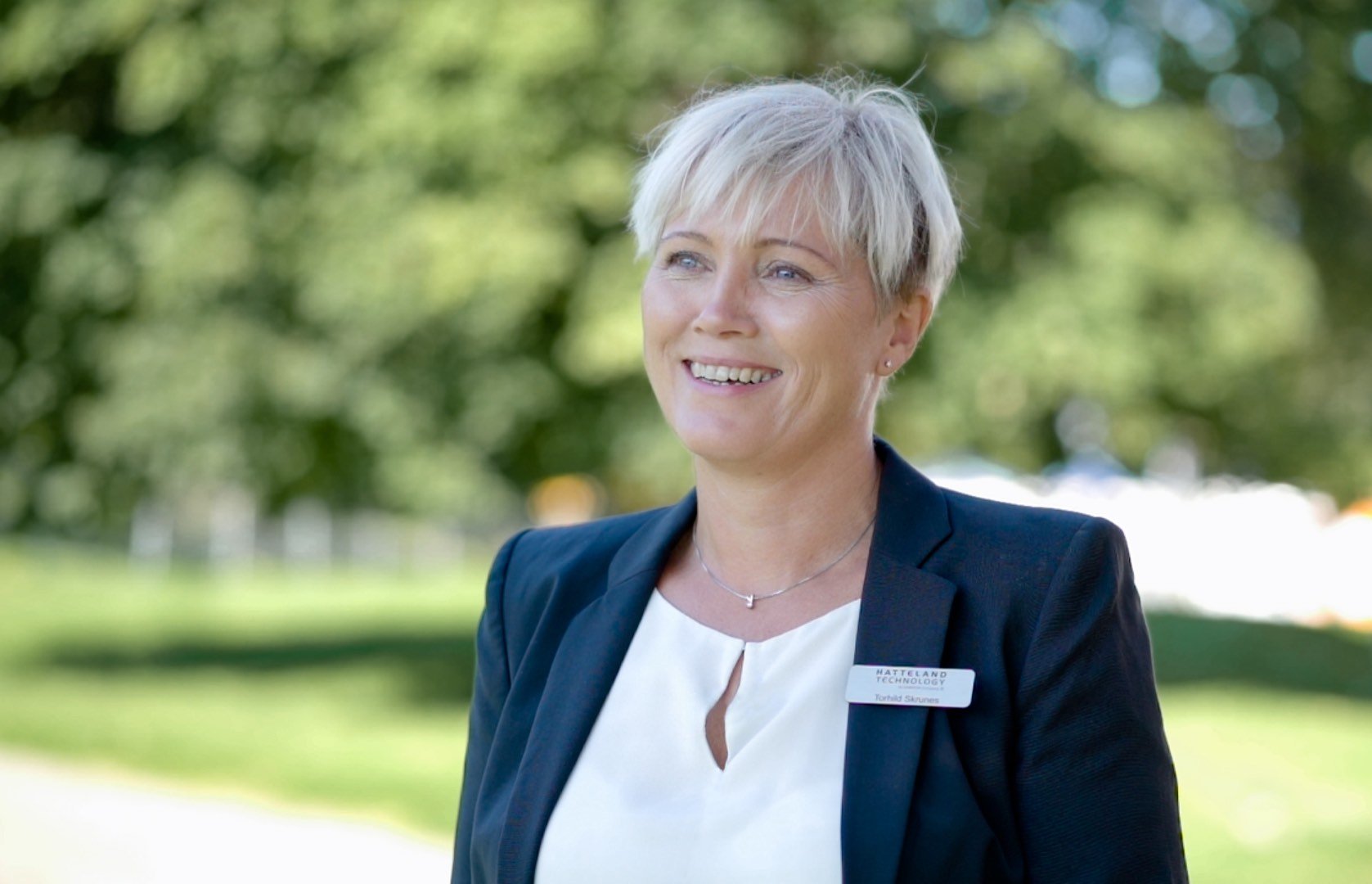 Ms. Torhild Skrunes from Hatteland Technology is in the centre of the photo, smiling. She is in the foreground, and the background is filled with summer green trees. This is outside of the convention hall in Trondheim, where Nor-Fishing 2022 took place. 