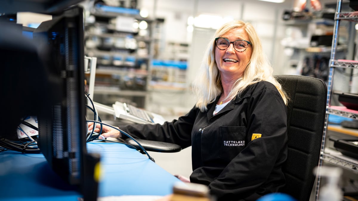 A staff member at Hatteland Technology is sitting down in front a computer. She is smiling. Wears glasses. 