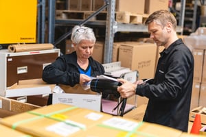 This image shows two employees discussing shipments at the Hatteland Technology warehouse in Aksdal, Norway. They are standing in the middle of lots of branded boxes, reviewing the shipment details. To the left, a woman, to the right, a man. They seem to be working in logistics. They are wearing black Hatteland Technology clothing.