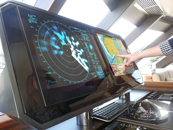 On the bridge of a ship or vessel. A finger is pointing at a nautical chart on a Hatteland Technology navigation monitor. A sonar display is also visible.