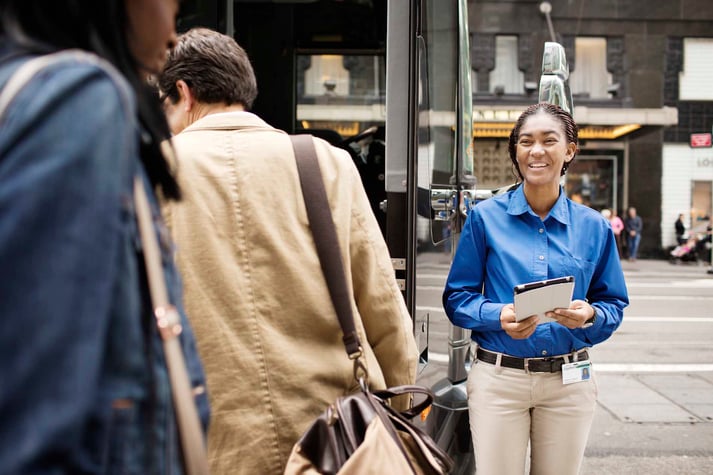 A smiling woman helps passenger get on board a bus. She uses a tablet for passenger servicing and management.