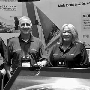 The USA team of Hatteland Technology: Scott Lenz, David Neal, and Donna Pallonetti. They are smiling, facing the camera. Behind them is some Hatteland Technology marketing material.