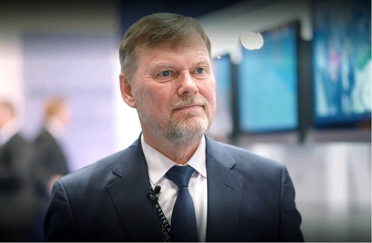 The picture shows Captain Jan Hansen at Nor-Shipping 2022. He is wearing a navy blue suit and tie, white shirt. In the background, an array of Hatteland Technology monitors mounted on the wall. 