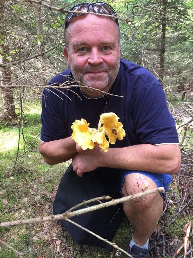 Maritime surveillance expert Anders Bjerketveit is holding up a bunch of chantarelles that he has picked from the forest. He is wearing blue clothes, and has sunglasses on his head. He is smiling. 