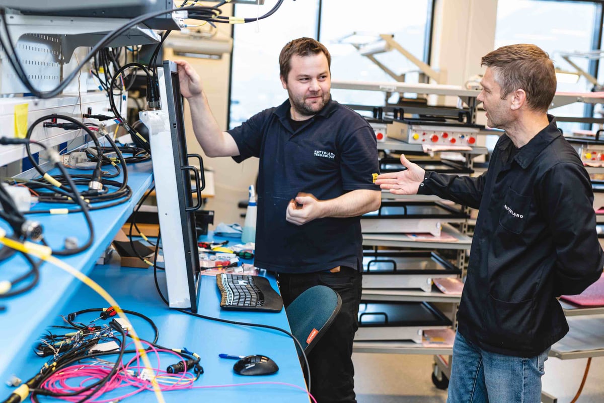 Two workers, wearing black Hatteland Technology shirts, are discussing something. One of them rests his hand on a monitor. There are lots of wires and technical parts around them. 