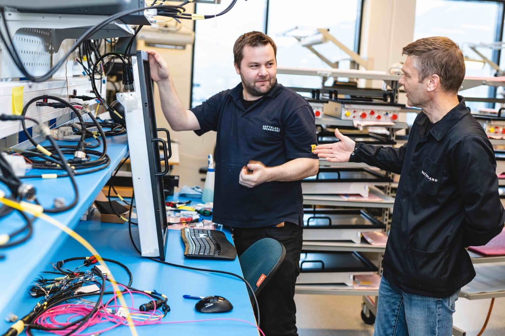 Two Hatteland Technology workers, both men, discuss a technical solution while manufacturing a piece of industrial IT hardware. One of them is holding a monitor.