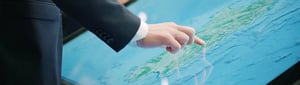 A hand is operating an electronic chart table from Hatteland Technology. The screen is displaying a map with topography. An island is visible, surrounded by blue water.