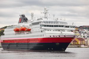 One of Hurtigruten's ships is departing or approaching port. It is a grey, calm day, with a small town visible in the background. The ship is black, red and white. We see the ship at a slight angle, head on.