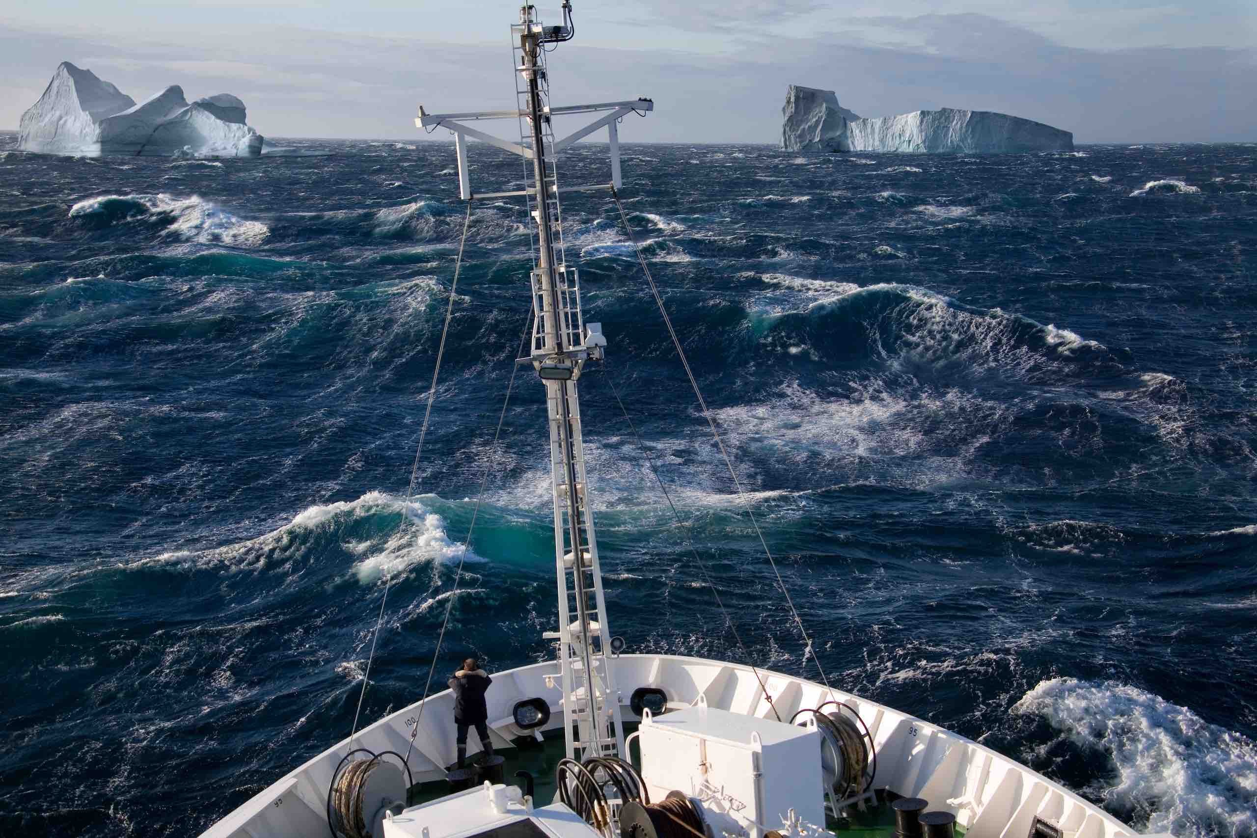 Illustrating the need for ruggedised IT equipment, the image shows the bow of a ship, facing stormy waters. Two icebergs are visible up ahead. The antenna mast is in the centre of the picture. The sea is dark blue, the ship is painted white. 