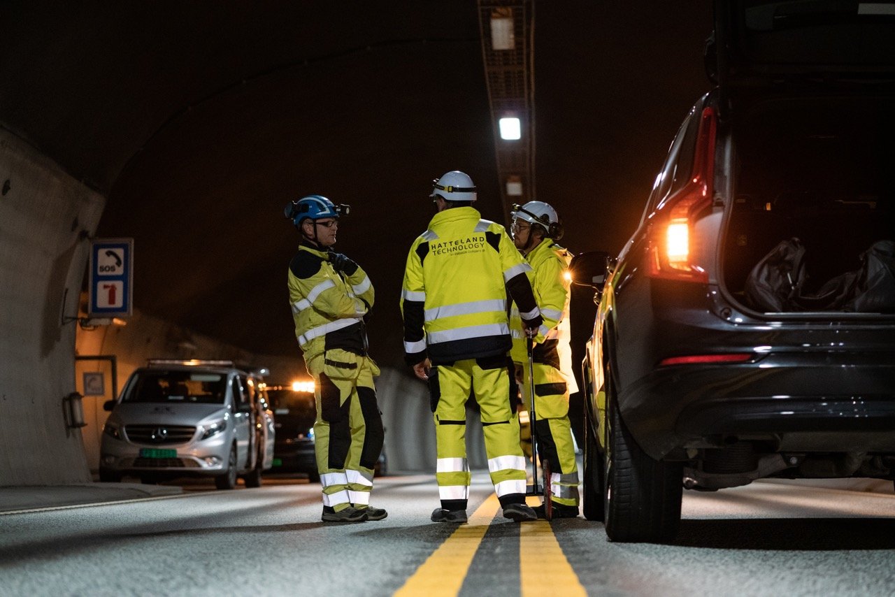 Three engineers discuss a surveillance solution in a road tunnel in Norway. They are wearing yellow, reflective work attire, helmets. The tunnel is dark. Two yellow centre lines run through the middle of the picture.