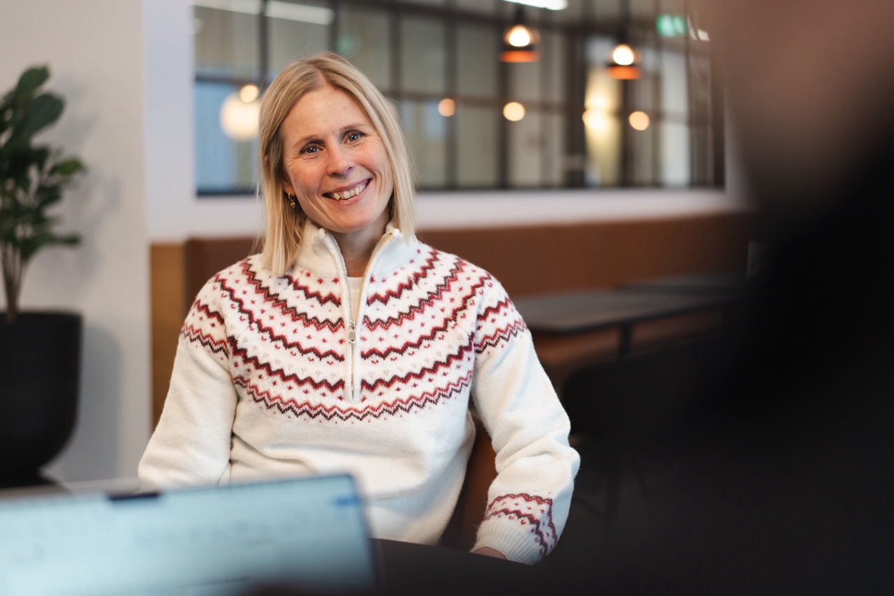 A picture of Anna Aulie, the Marketing & Communications Manager at Hatteland Technology. She is smiling, looking directly at the camera, and wearing a knitted sweater. There are lamps and a brown sofa in the background.