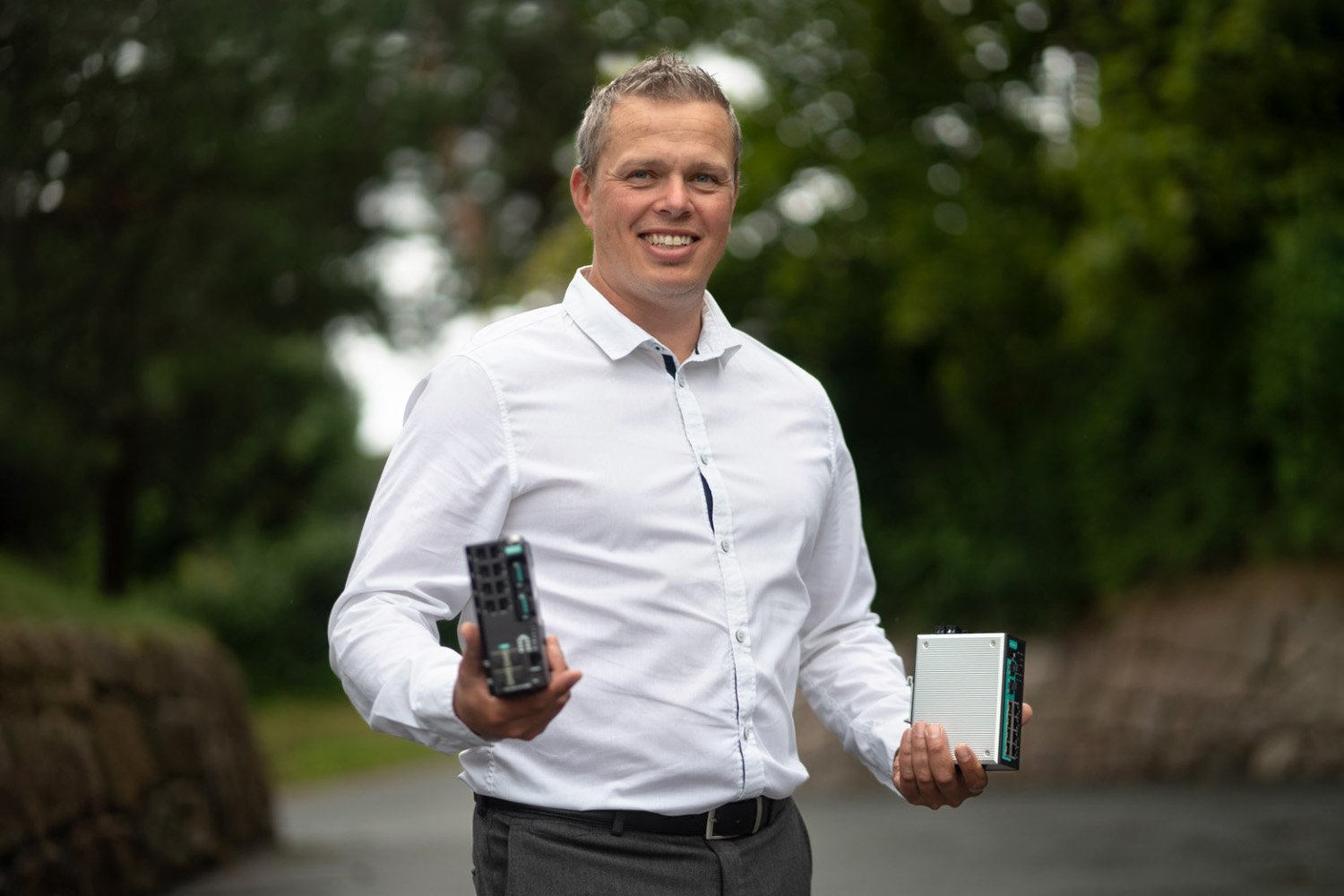 The picture shows Mr. Gøran Labrå, Business development director at Hatteland Technology, holding two Moxa switches, one of which is the EDS-G4000 series. He is wearing a white shirt and is smiling. There are green trees in the background.