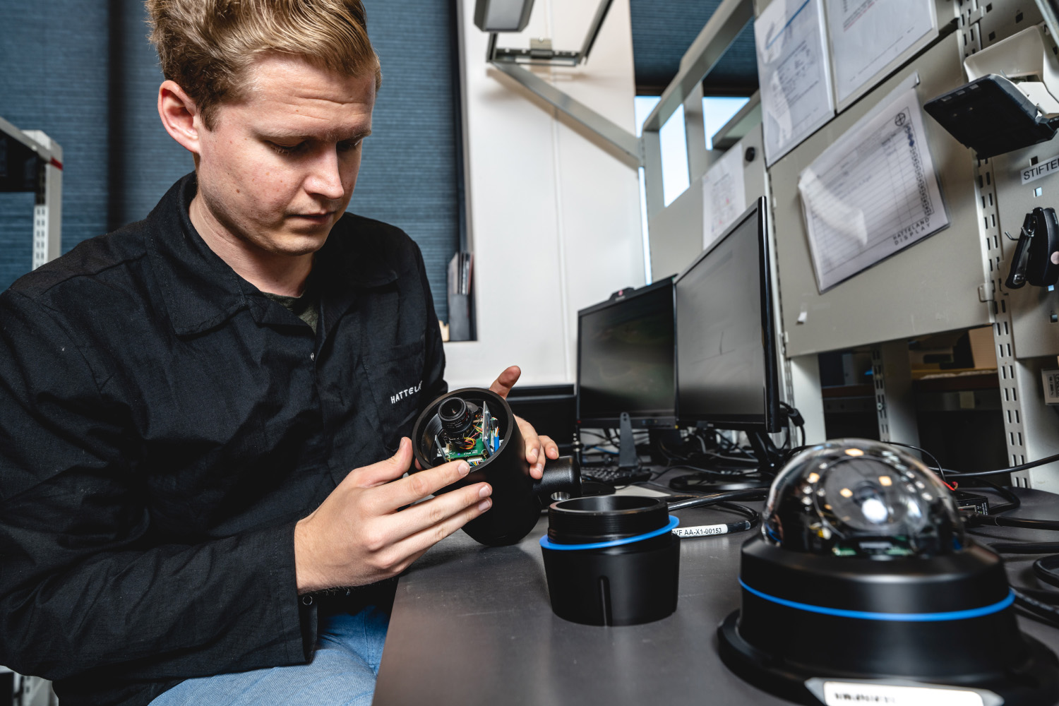 A Hatteland Technology technician is assembling a SeaHawk bullet camera, a rugged CCTV camera for maritime applications. He is wearing a black shirt and blue jeans. Even closer to the camera is a Hatteland Technology SeaHawk dome camera.