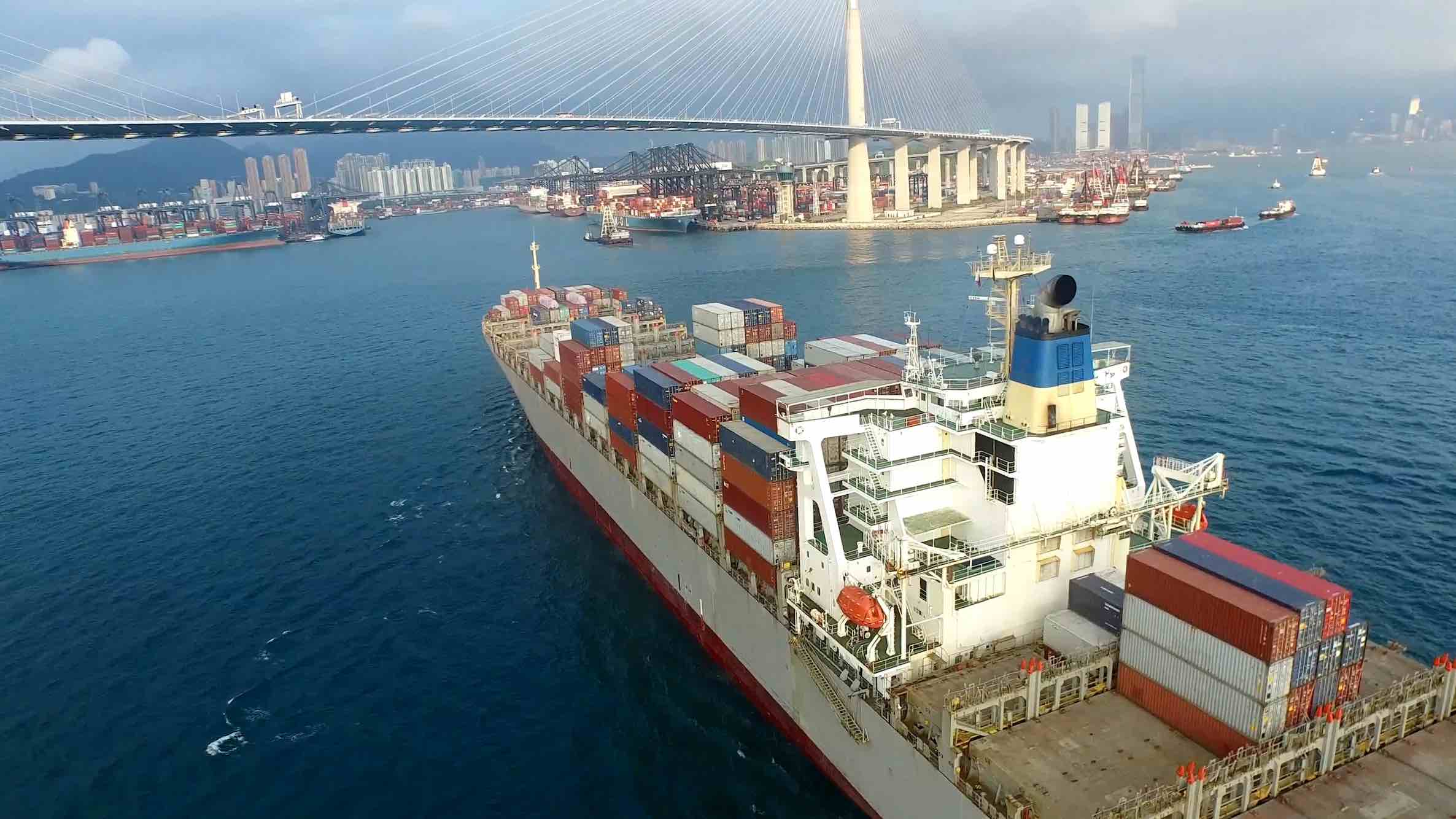 Aerial view of a loaded, white cargo ship that's about to dock in a busy city port. Straight ahead is a suspended bridge. Many others ships are nearby. 