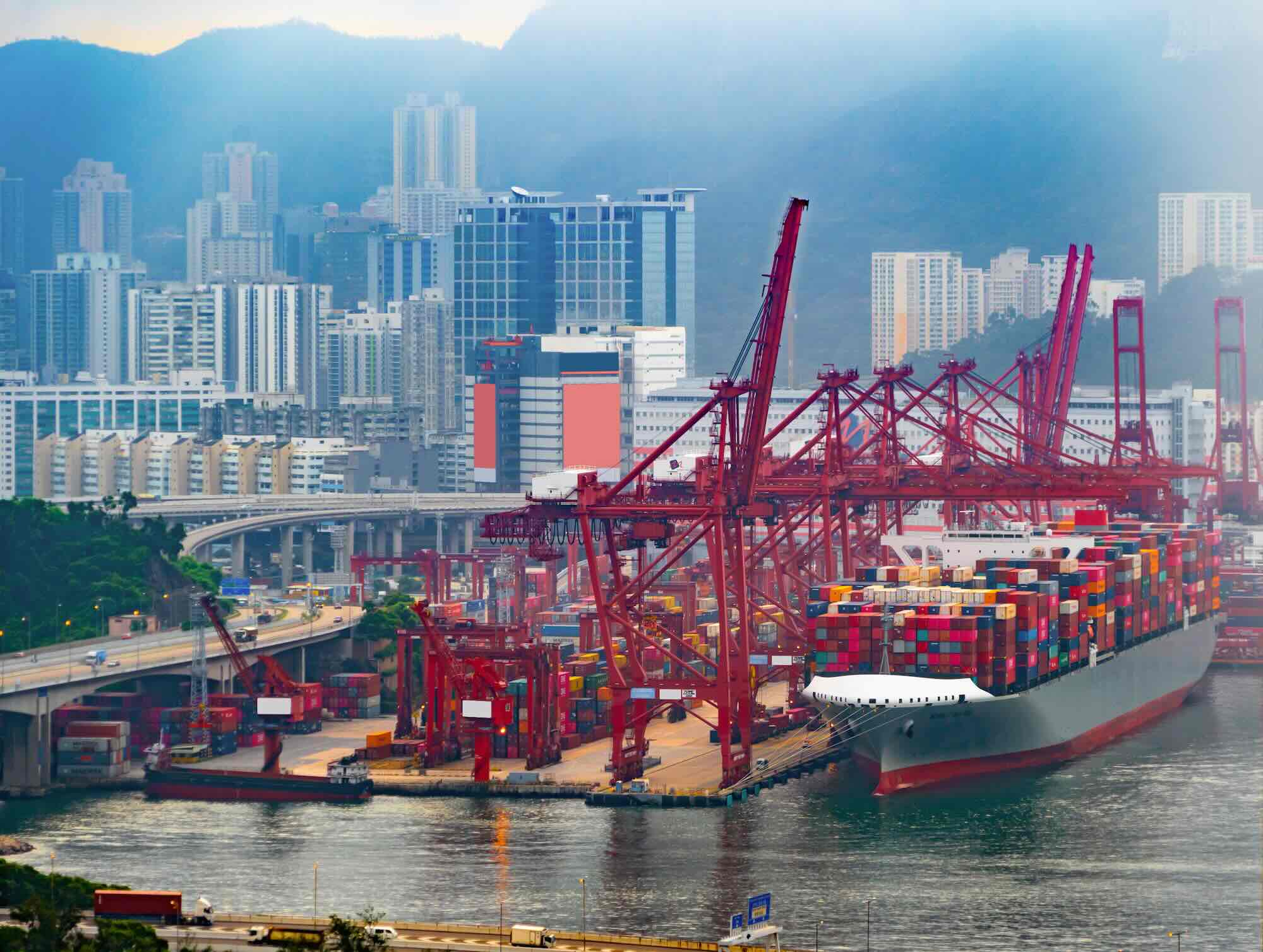 Aerial view of a cargo ship about to dock in a shipyard or dock area. There are large, red cranes in the middle of the picture. In the background there is a city. The photo illustrates the web of possible connections and vulnerabilities ships face with respect to digitalization. Maritime cyber security relates to both ship side and land side operations.